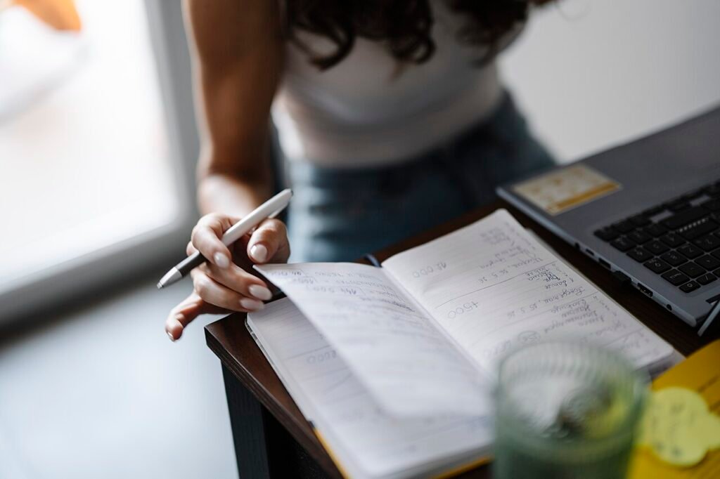 Persona sentada a la mesa tomando notas en un cuaderno, junto a una computadora portátil